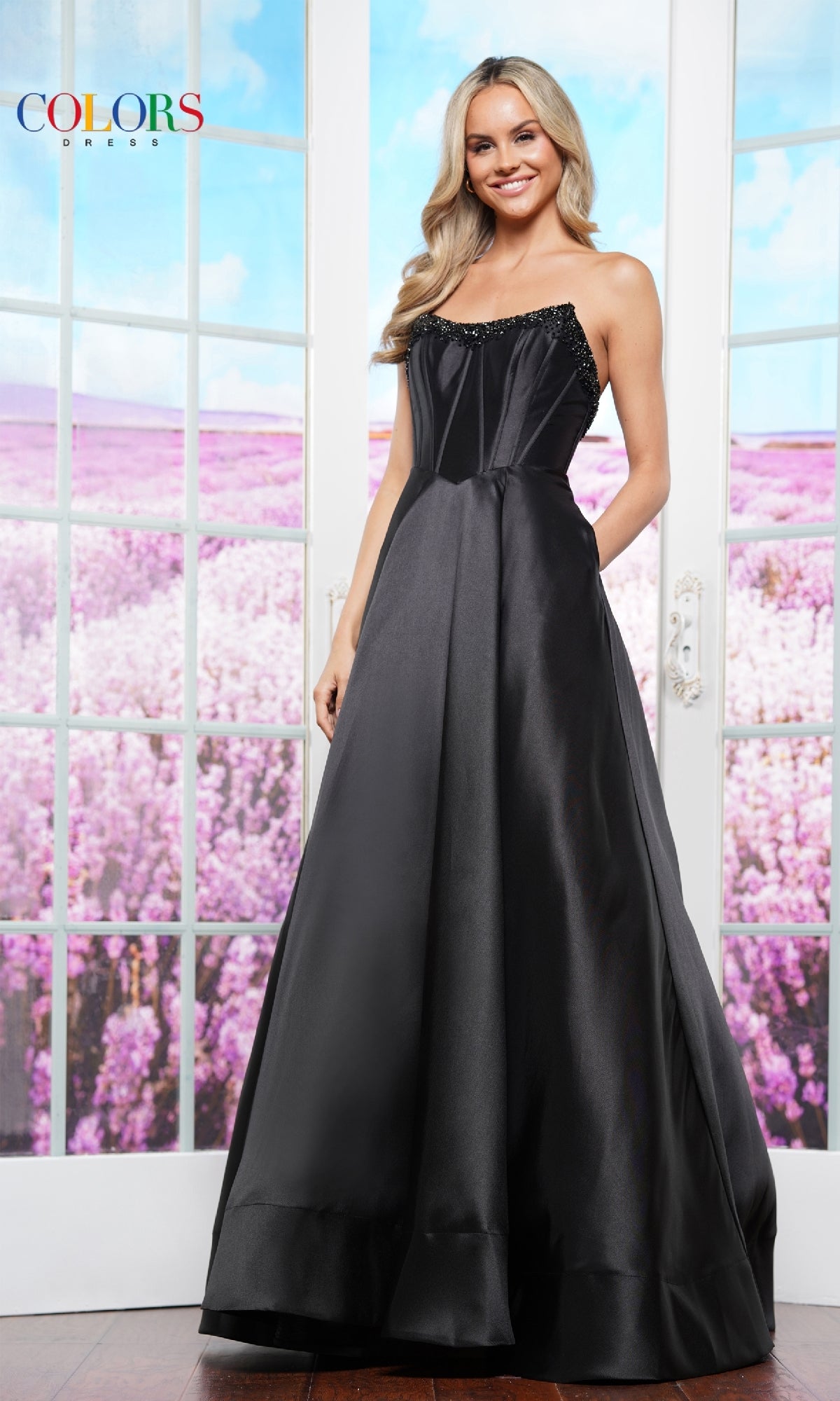 A woman wearing the Strapless Corset Prom Ball Gown: Colors Dress 3478 stands indoors before glass doors, showcasing a stunning field of purple flowers outside.